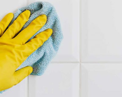 A gloved hand using a cloth to clean white bathroom tiles.